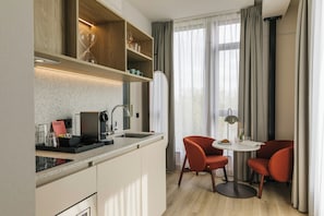 Een compacte keuken met een ingebouwde koffiezetapparaat, een kleine eettafel met twee stoelen, en een grote spiegel boven het aanrecht.