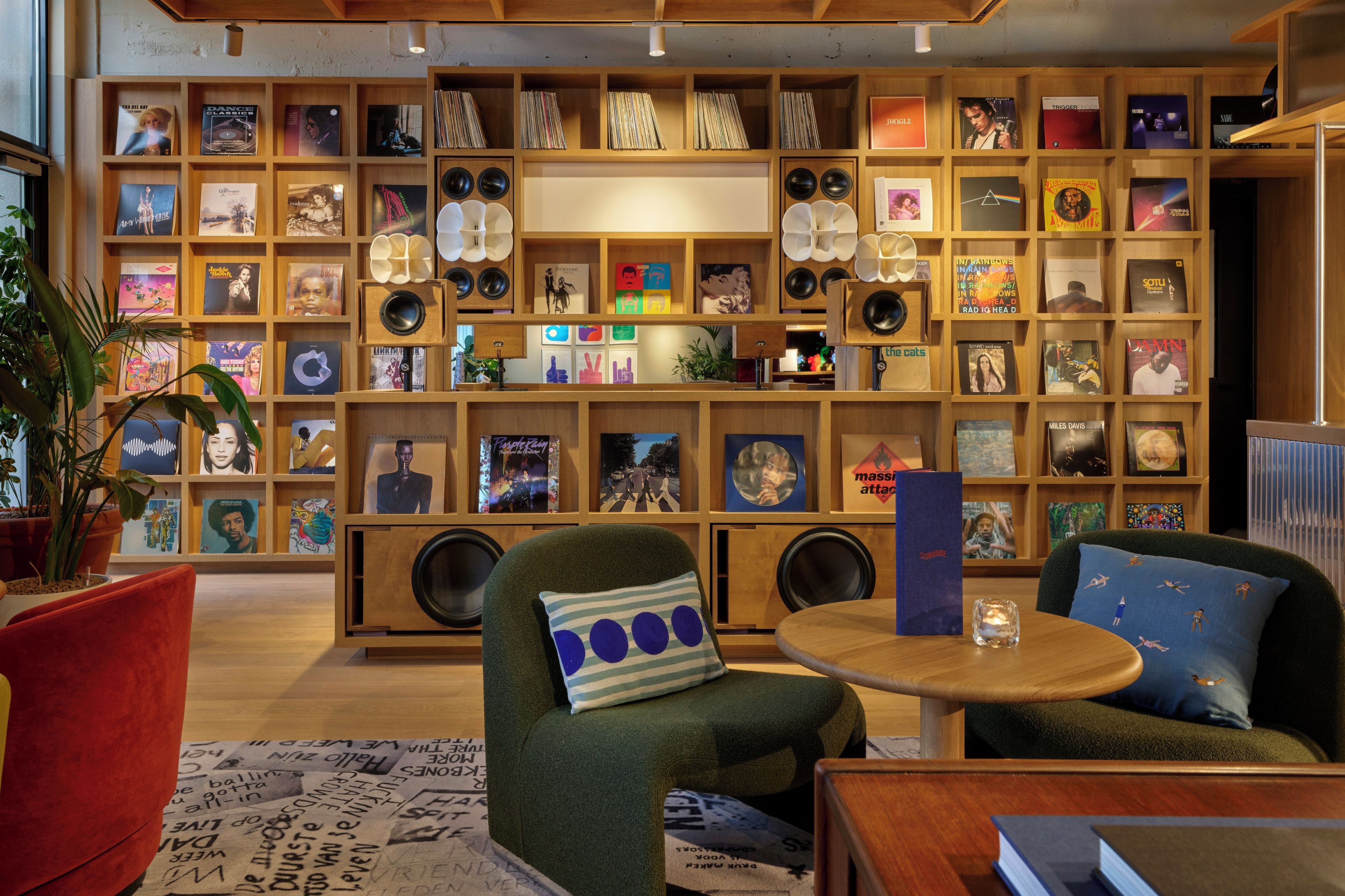 Une pièce avec une bibliothèque remplie de disques vinyles, un fauteuil vert, une table ronde en bois et un canapé rouge.