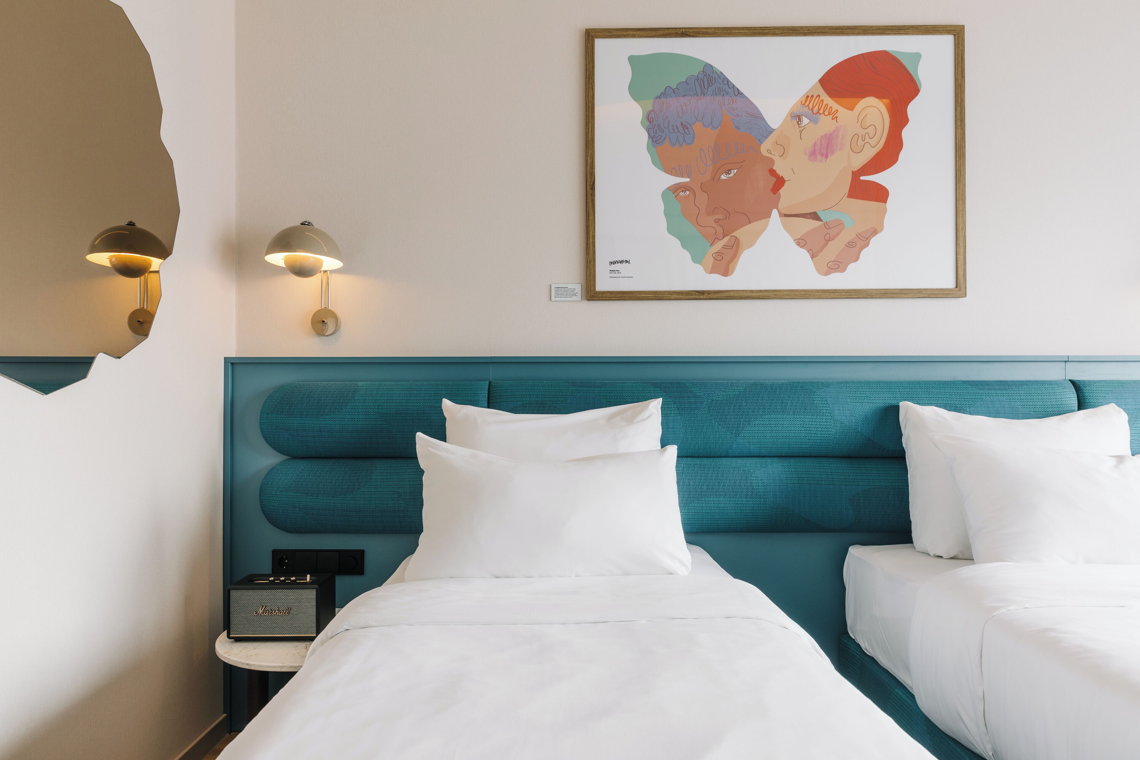 A hotel room with a teal headboard, white bedding, a framed artwork on the wall, and a bedside table with a lamp.