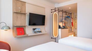 A hotel room with a bed, a TV mounted on the wall, a shelf with a red book, and a clothing rack with a striped shirt.
