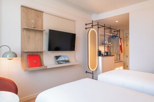Une chambre d’hôtel avec un lit, un téléviseur fixé au mur, une étagère avec un livre rouge et un portant à vêtements avec un t-shirt rayé.