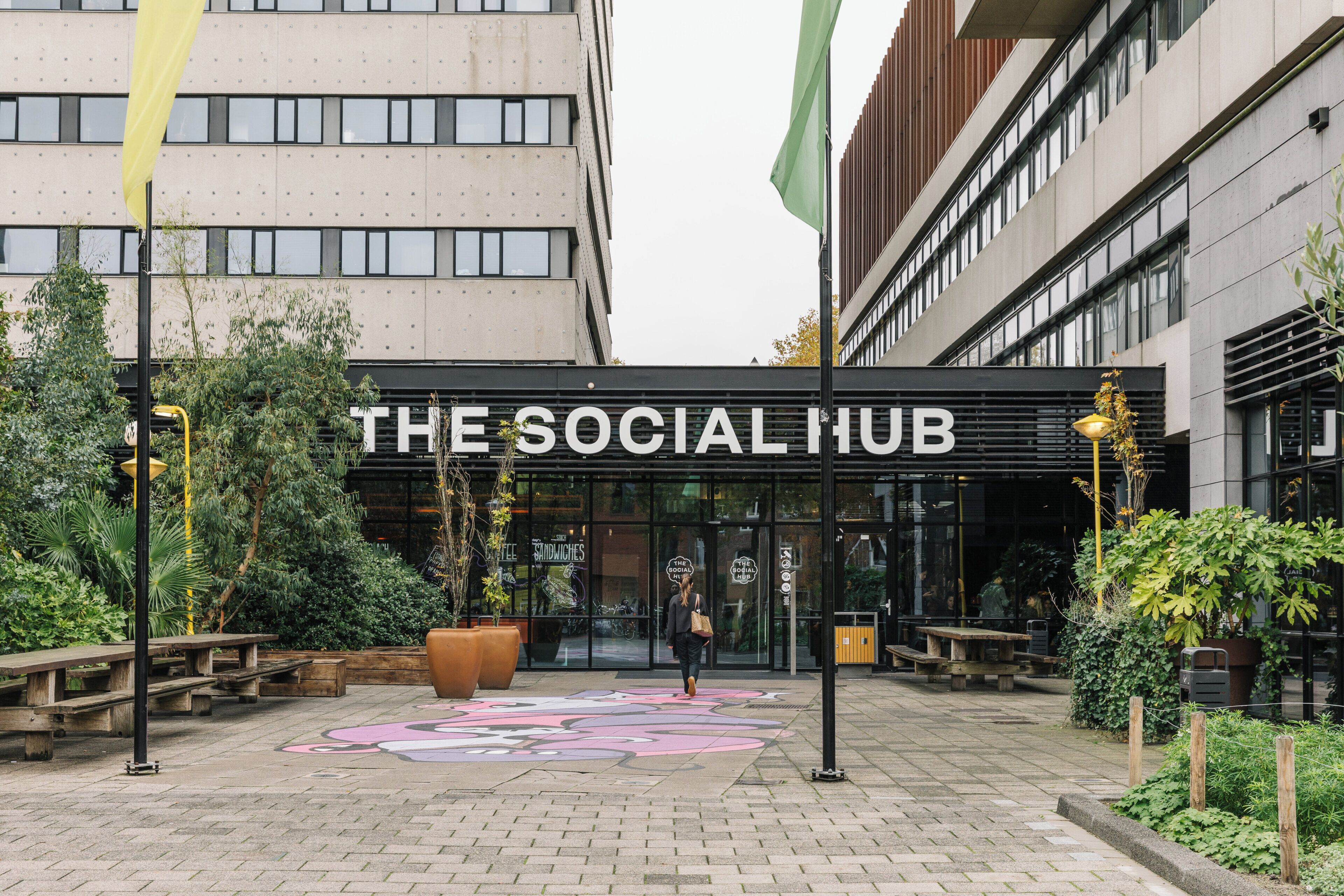 A building entrance with a sign reading 'The Social Hub,' surrounded by outdoor seating and greenery.