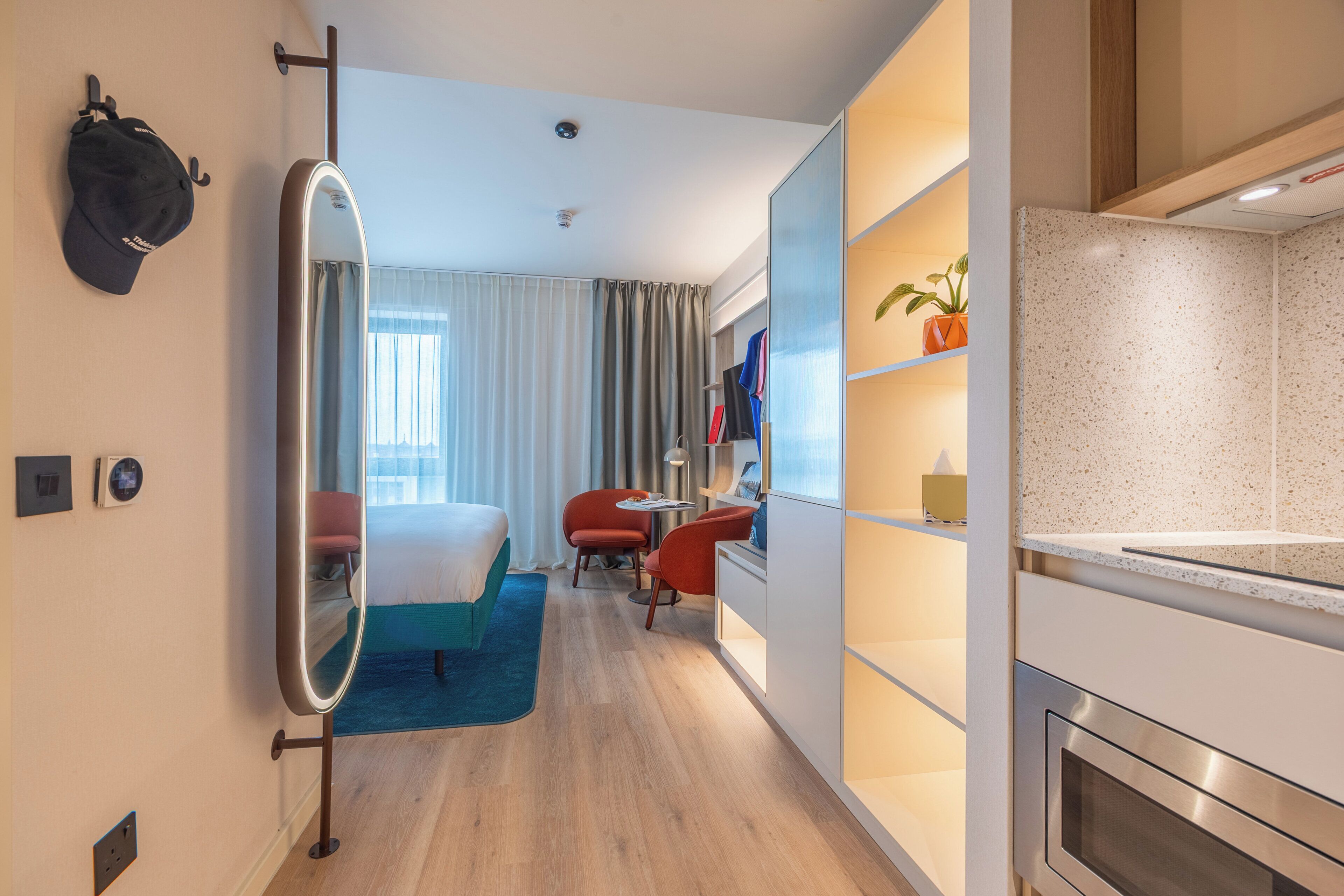 Et moderne hotelværelse med seng, skrivebord, stol, spejl, lille bord og tekøkken.
