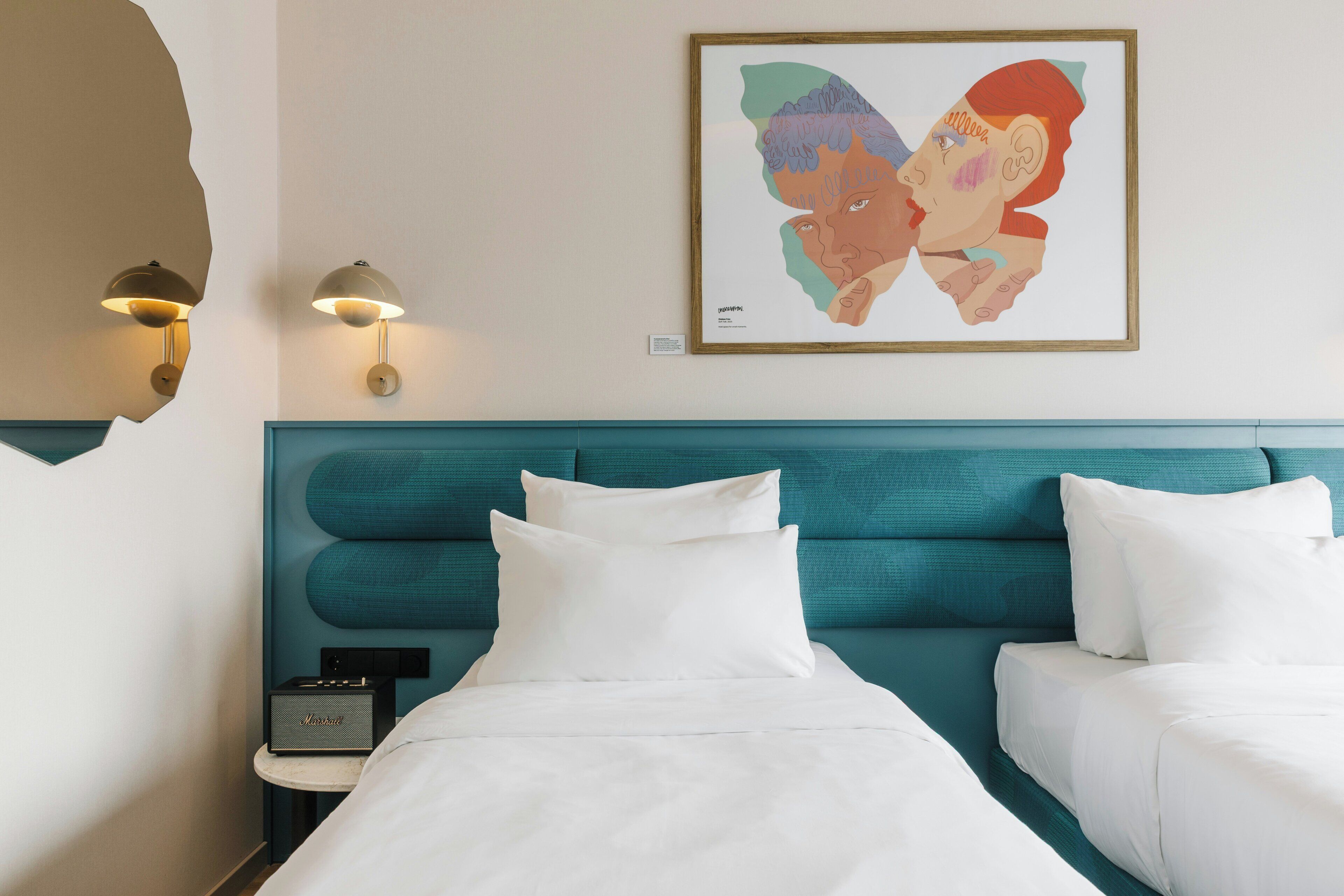 A hotel room with a teal headboard, white bedding, a framed artwork on the wall, and a nightstand with a lamp.