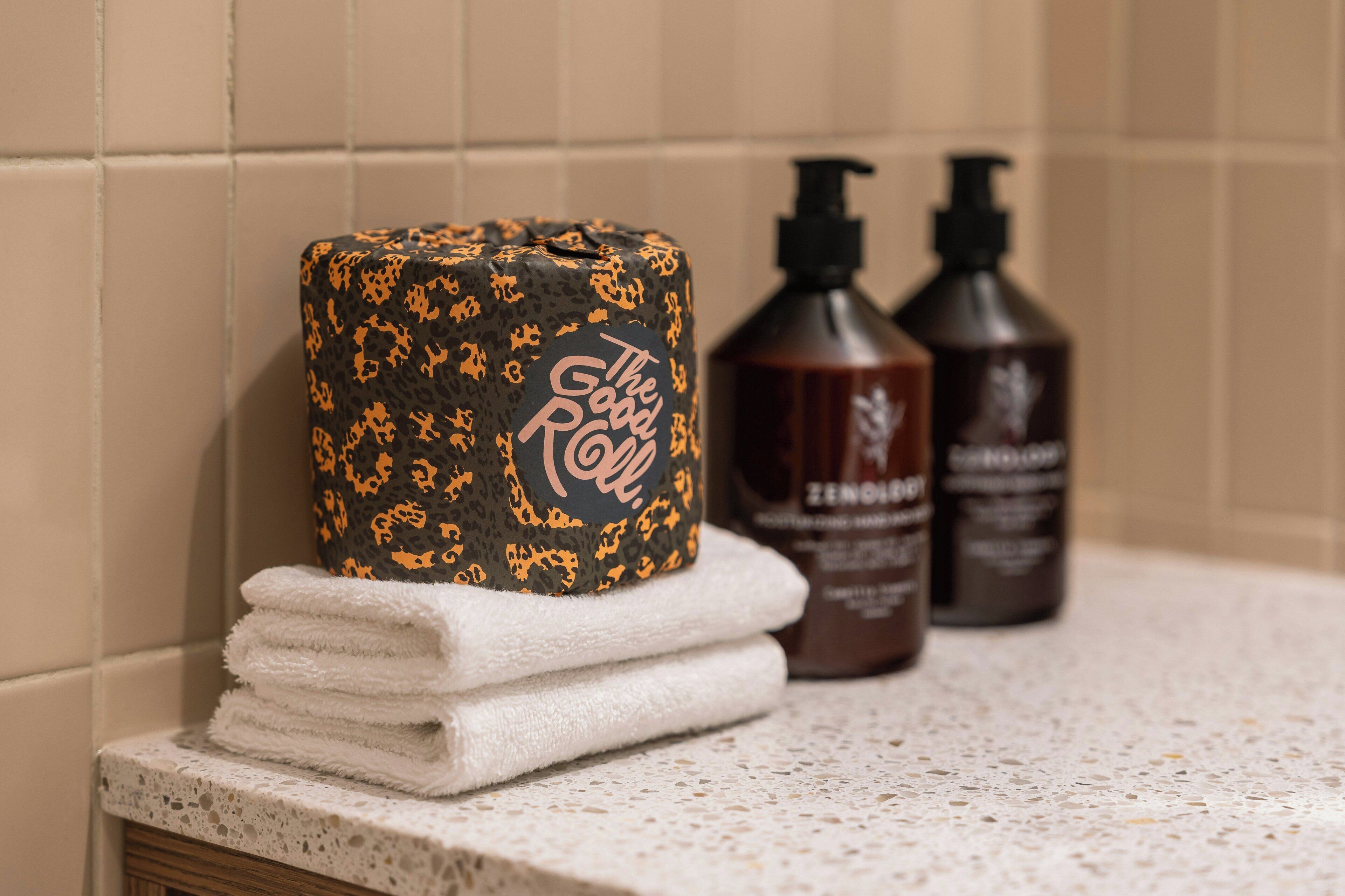 A bathroom with a toilet paper roll, two bottles of shampoo, and a stack of white towels.