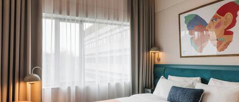 A hotel room with a bed, a chair, a nightstand, a lamp, and a window with curtains.
