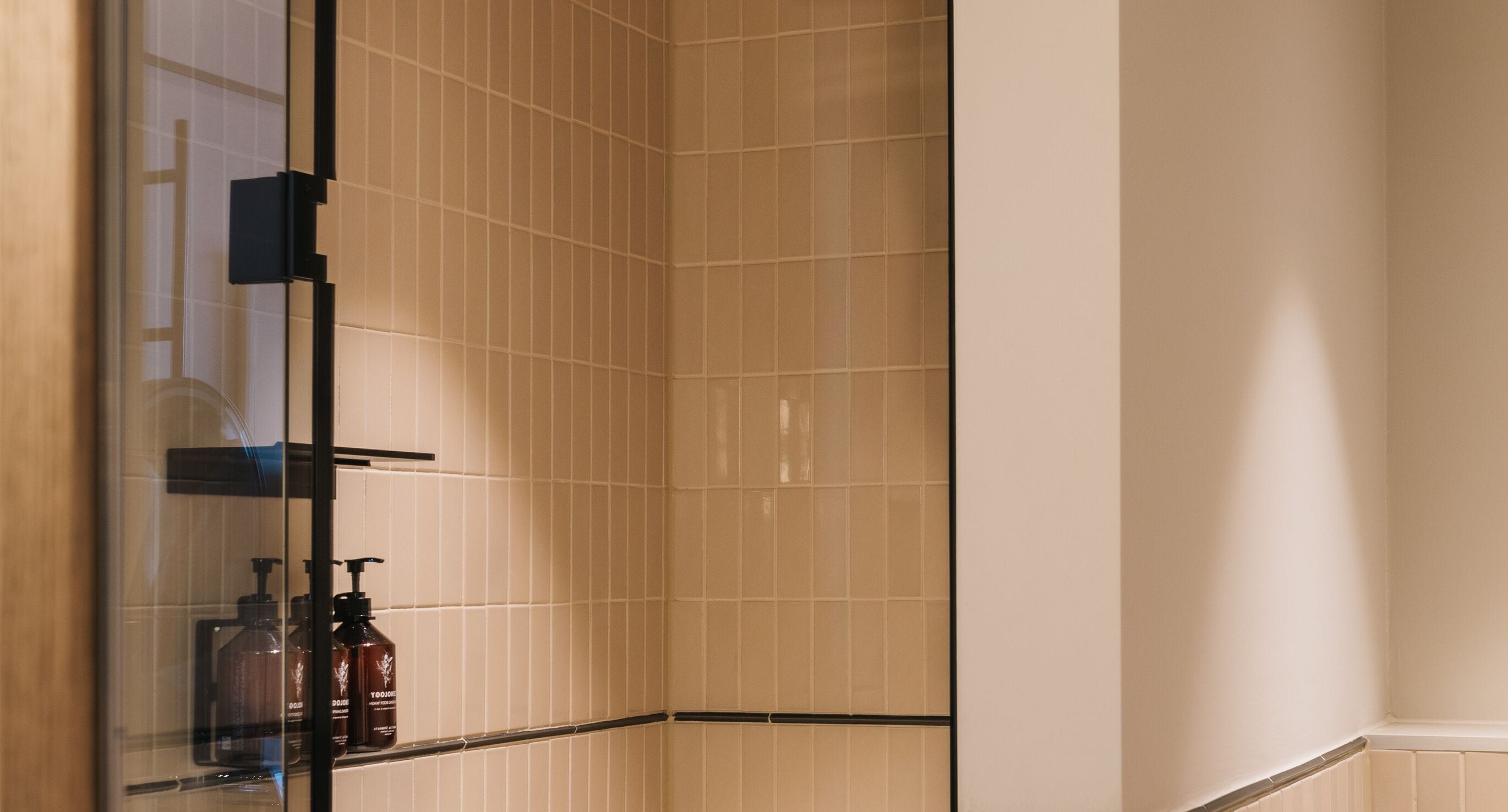A corner of a bathroom with beige tiles, a glass shower enclosure, and a shelf with toiletries.