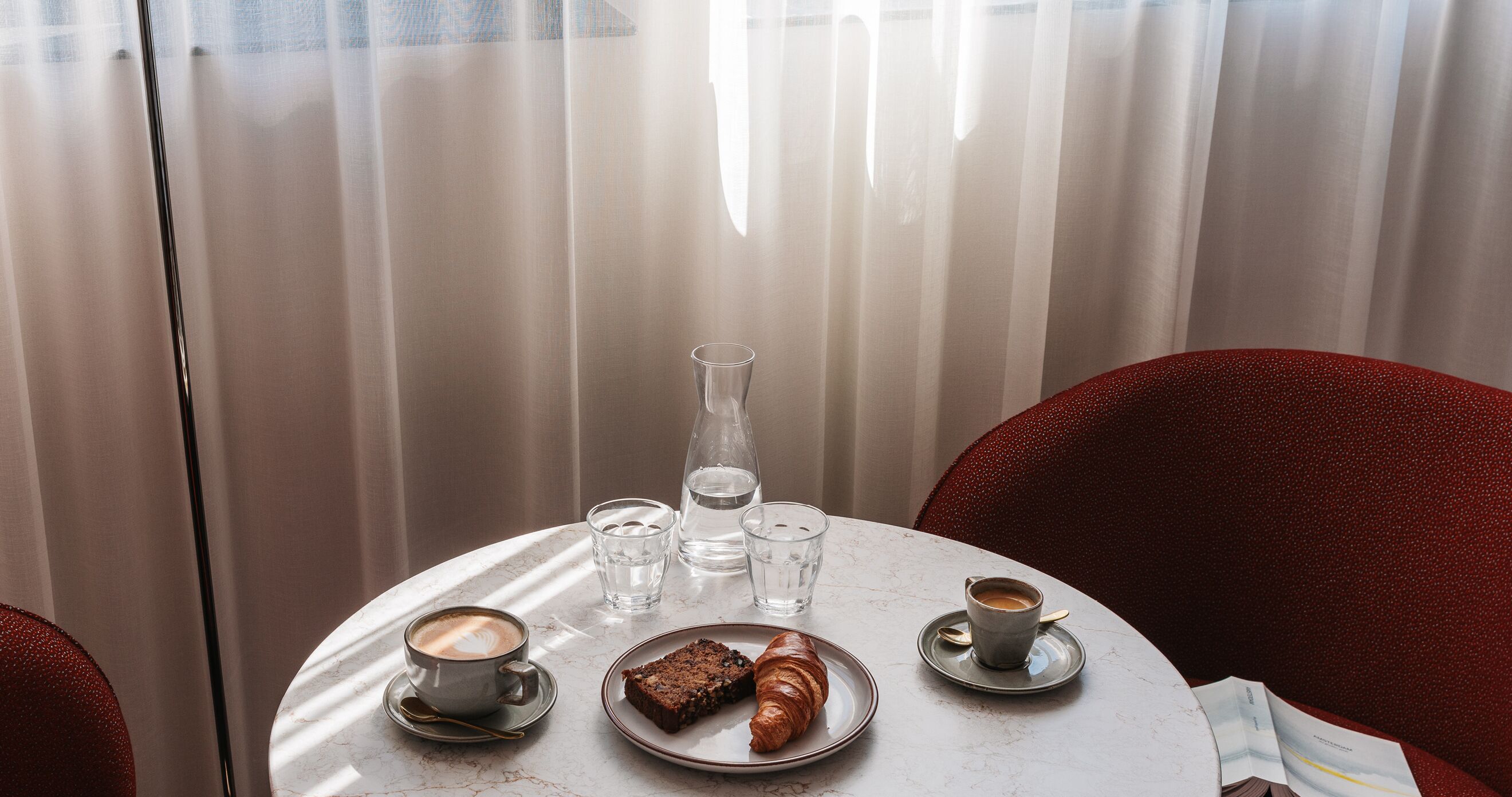 Een ronde tafel met een croissant, een glas water, een glas ijswater en een kop koffie.