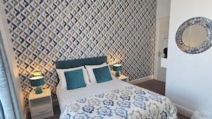 Une chambre avec un mur à motifs, un lit recouvert d’une couvre-lit à fleurs, deux lampes de chevet et un miroir rond accroché au mur.