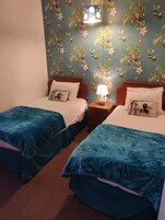 Habitación con dos camas, cada una con una colcha azul y un almohadón blanco. Entre las camas, un mesita de noche con una lámpara. La pared presenta un estampado de flores.