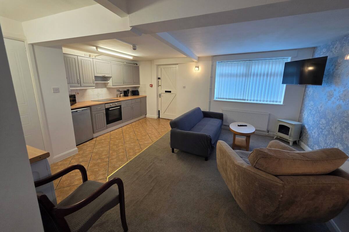 Een compacte leefruimte met een kitchenette, een bank, een tafeltje en een aan de muur bevestigde televisie.