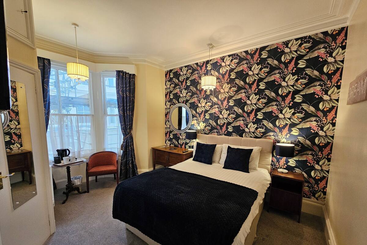 Een slaapkamer met een groot bed, een nachtkastje, een kaptafel, een stoel en een raam met gordijnen.