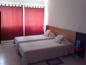 A hotel room with two single beds, a red curtain, a small table with a blue object, and a tiled floor.