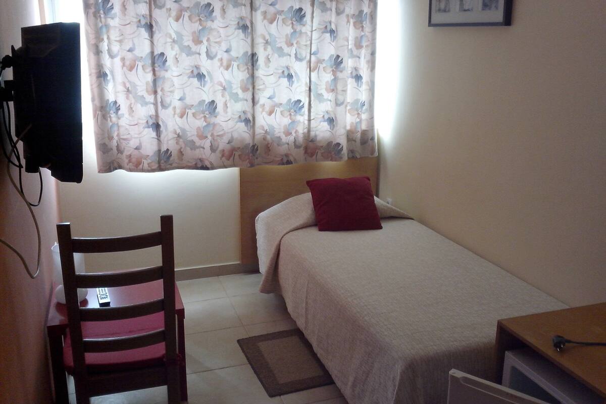 Un dormitorio pequeño y ordenado con una cama individual, una silla, un escritorio y un ventanal con cortinas florales.