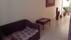 A living room with a dark patterned sofa, a small side table, and a decorative wall hanging. There are potted plants and a wooden sculpture on a tiled floor. - Tarrafal Residence (Tarrafal)