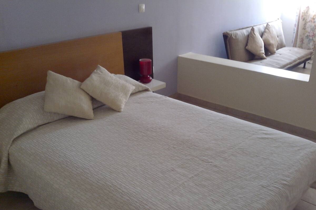 Una cama bien hecha con almohadas, una mesita de noche con un objeto rojo y un sofá con cojines.