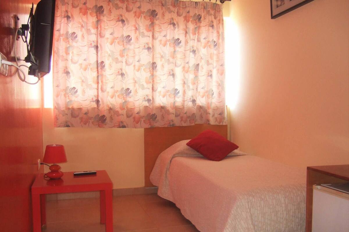 Una habitación pequeña y acogedora con una cama individual, una mesa roja con una lámpara y un televisor montado en la pared.