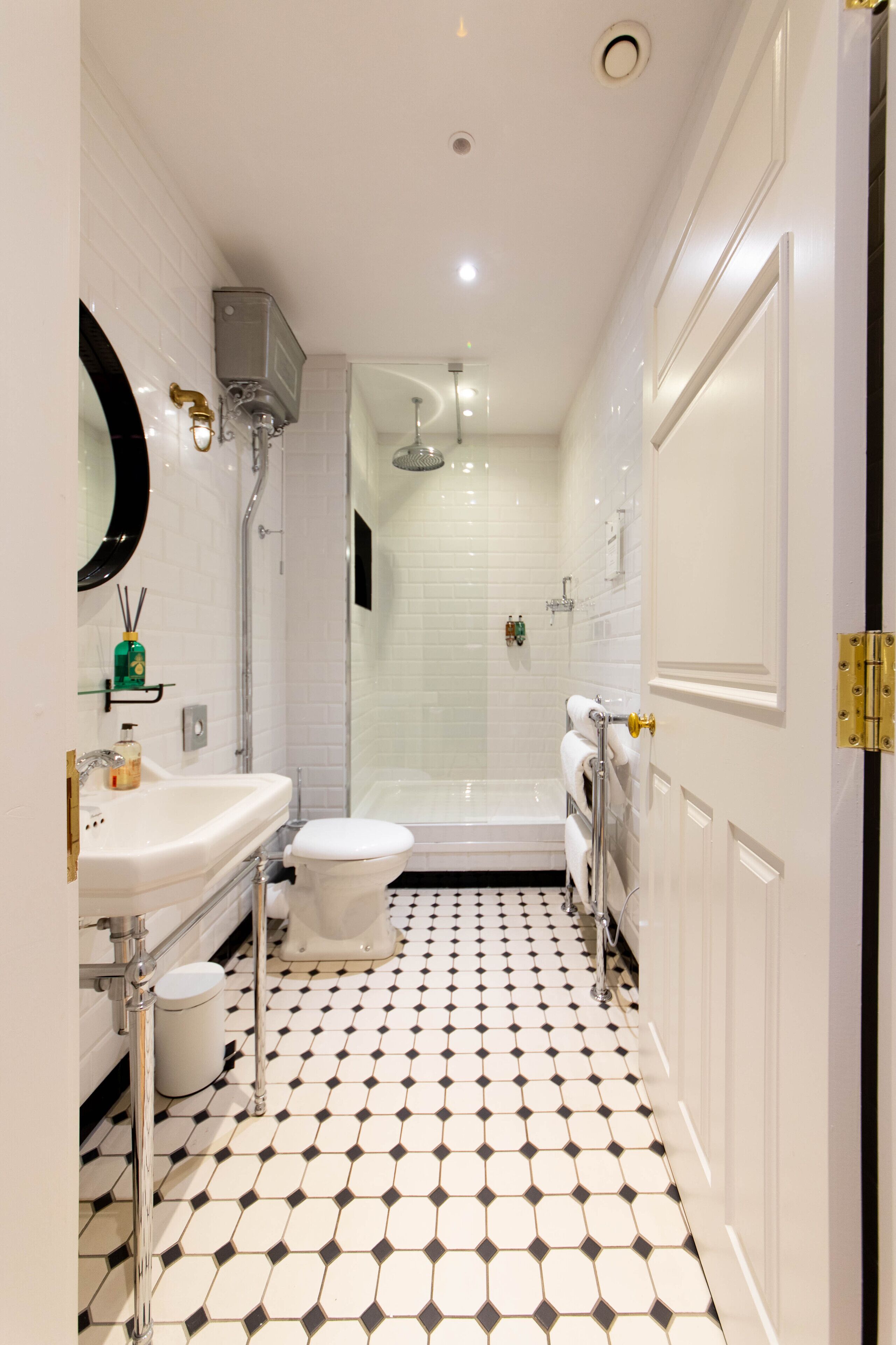 Une salle de bain avec un lavabo, des toilettes et une douche, le tout de couleur blanche. Le sol est recouvert d’un motif à carreaux noirs et blancs.