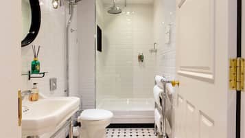 Une salle de bain avec un lavabo, des toilettes et une douche, le tout de couleur blanche. Le sol est recouvert d’un motif à carreaux noirs et blancs.