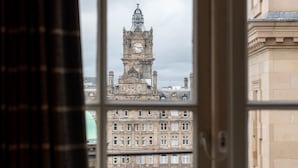 Une vue d’un bâtiment historique, avec un imposant clocher et un drapeau, que l’on aperçoit à travers une fenêtre ornée d’un rideau.
