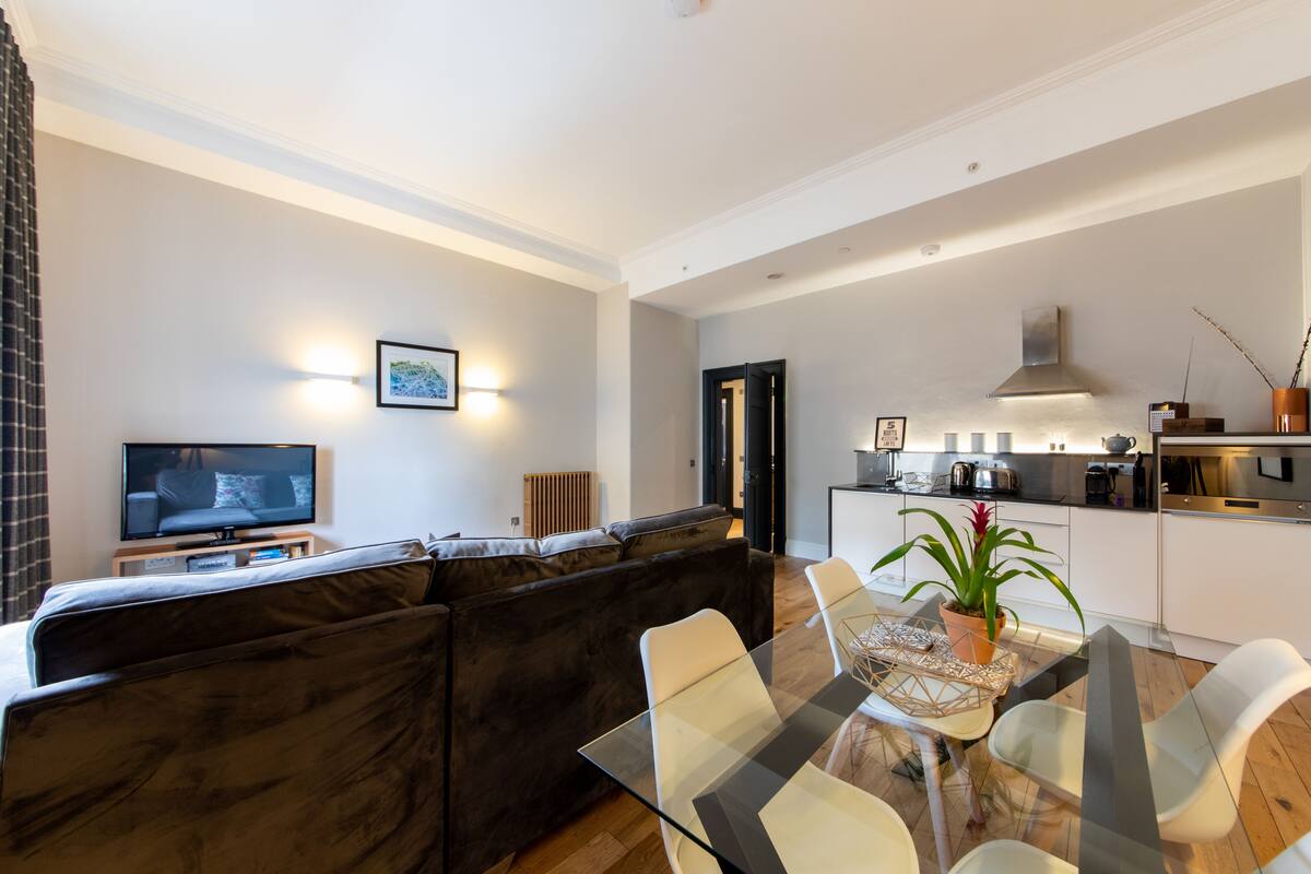 Un salon moderne avec un canapé foncé, une table à manger en verre et un coin cuisine équipé d’une cuisinière et de placards.