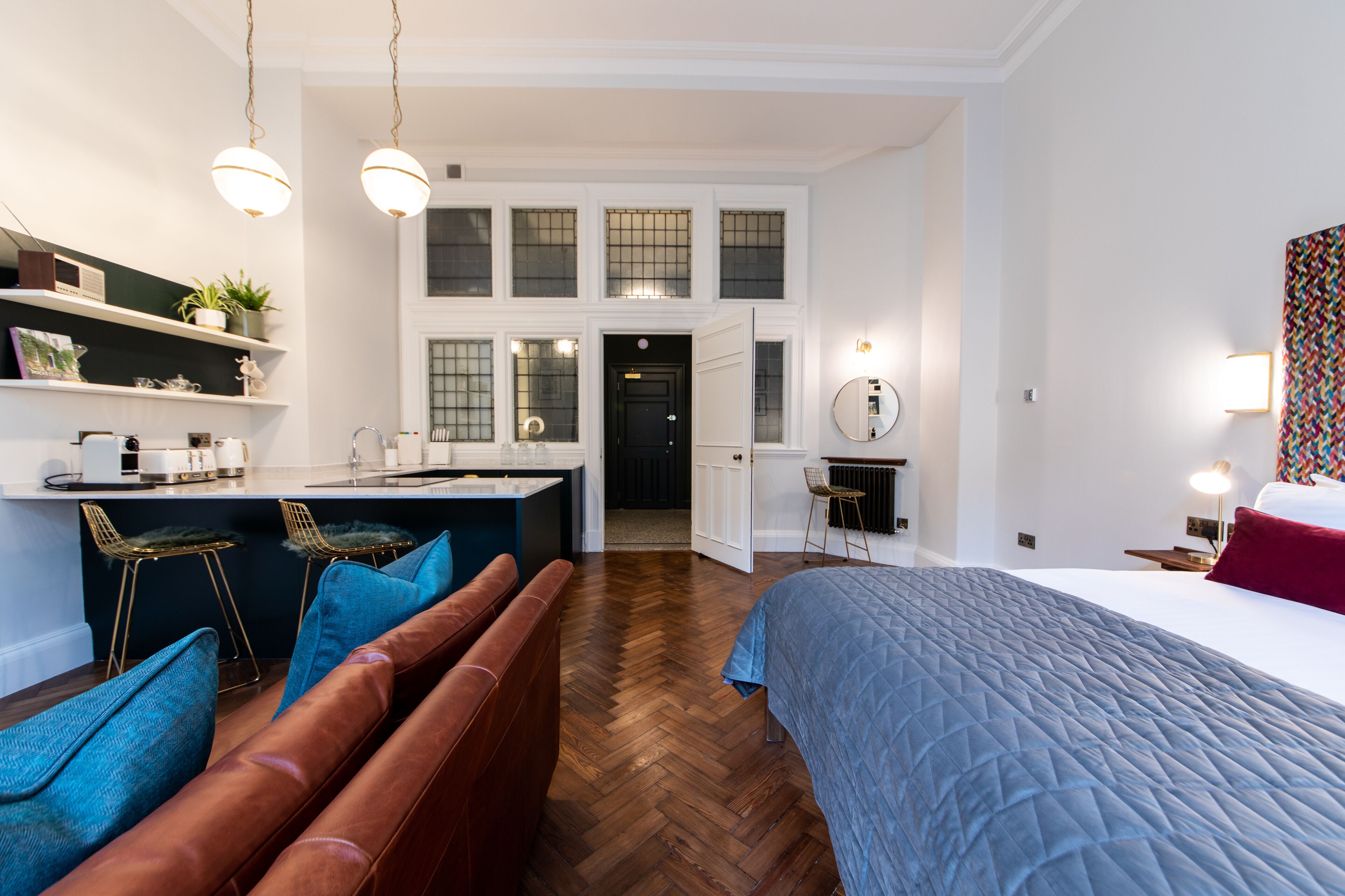 A modern hotel room with a large bed, a seating area with a brown sofa and blue cushions, a kitchenette with bar stools, and a view of the city through large windows.