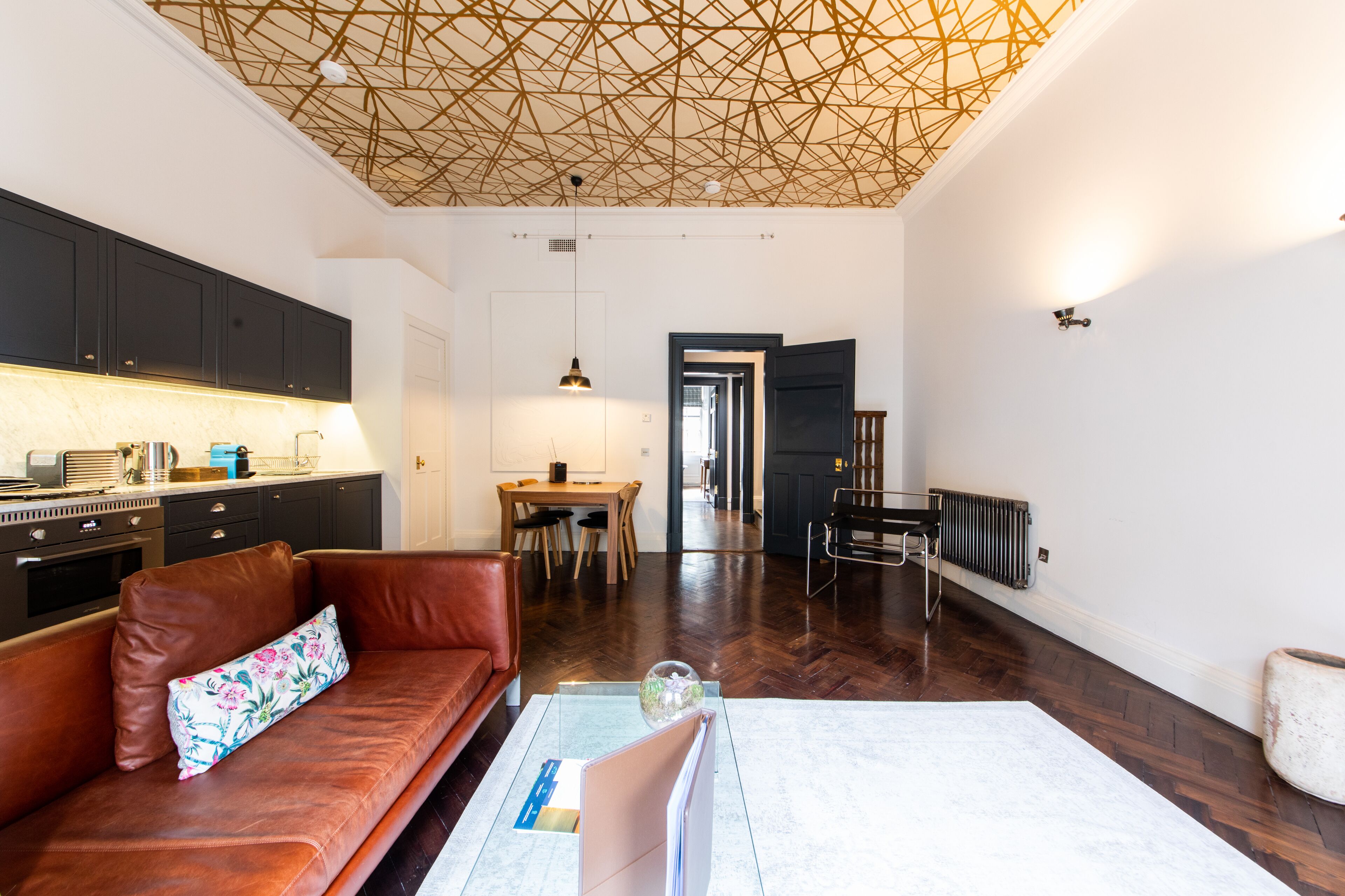Eine moderne Küche mit dunklen Schränken, ein Essbereich mit Holztisch und -stühlen sowie ein Wohnzimmer mit Ledersofa und Couchtisch.