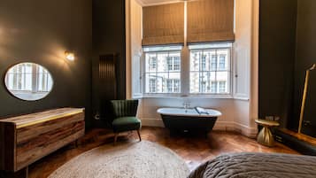 Une salle de bain moderne avec une baignoire sur pieds, un miroir rond, une chaise verte et une grande fenêtre donnant sur des immeubles.