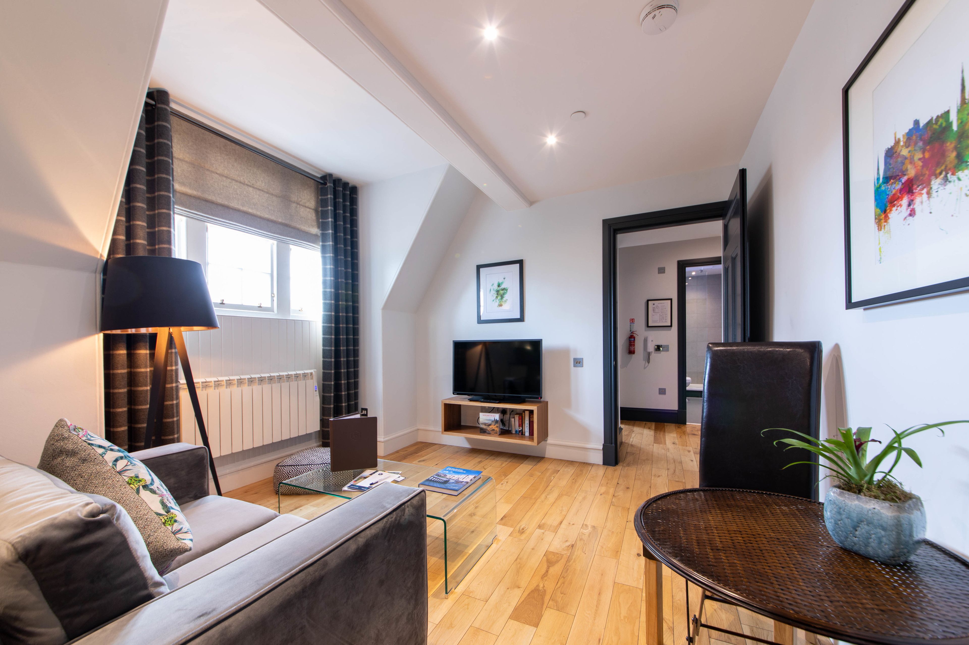 Una sala moderna con sofá, una mesa de centro, un televisor sobre un mueble bajo, una mesa de comedor con una planta y un ventanal con cortinas.