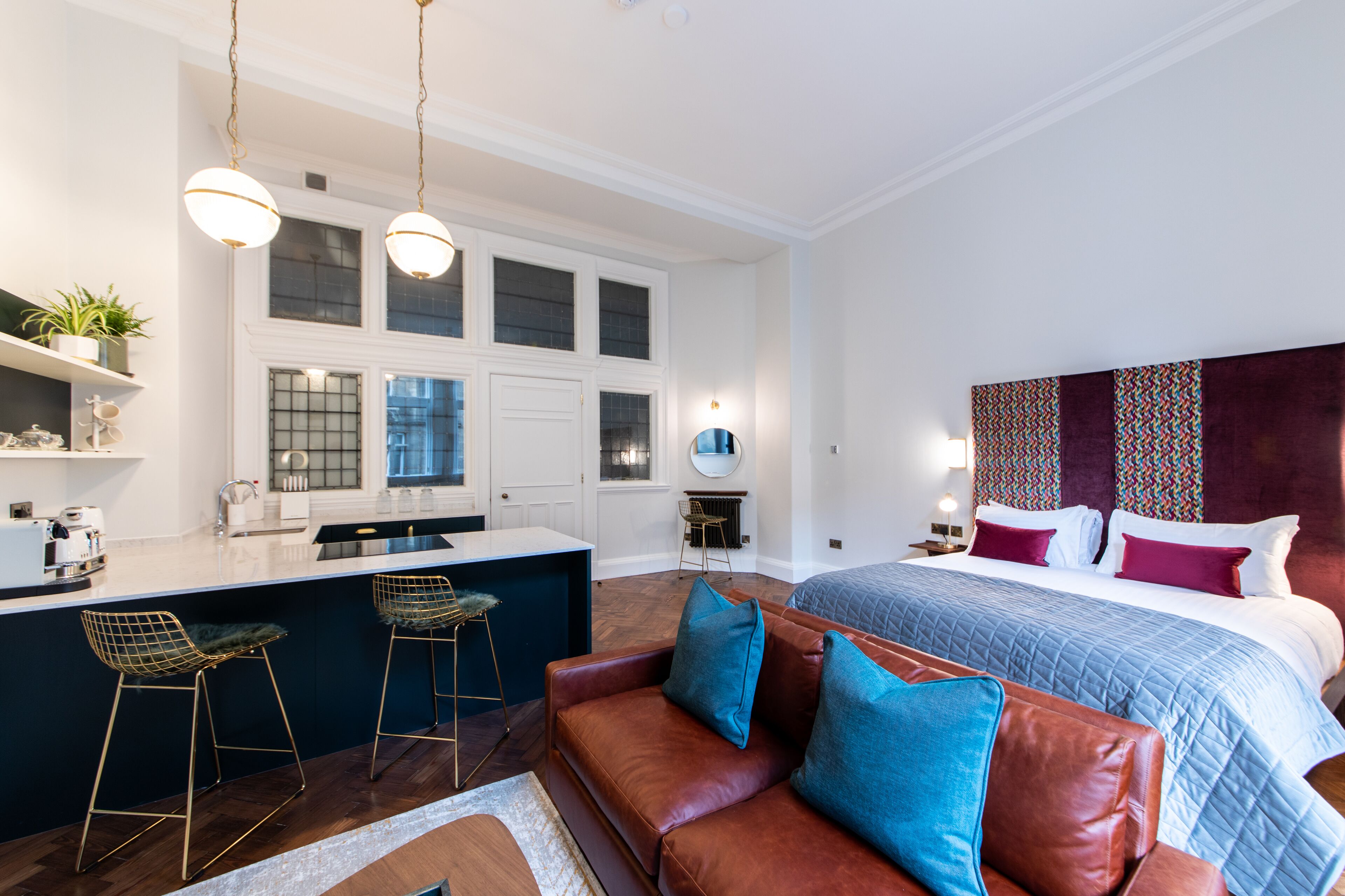 A modern hotel room with a large bed, a sitting area with a brown leather sofa, a kitchen area with a bar, and a large window.