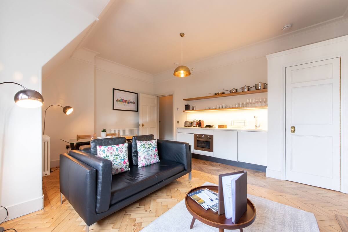 Un salon moderne avec un canapé en cuir noir, une table basse avec des magazines, un luminaire suspendu et un coin cuisine doté de meubles blancs et d’étagères en bois.