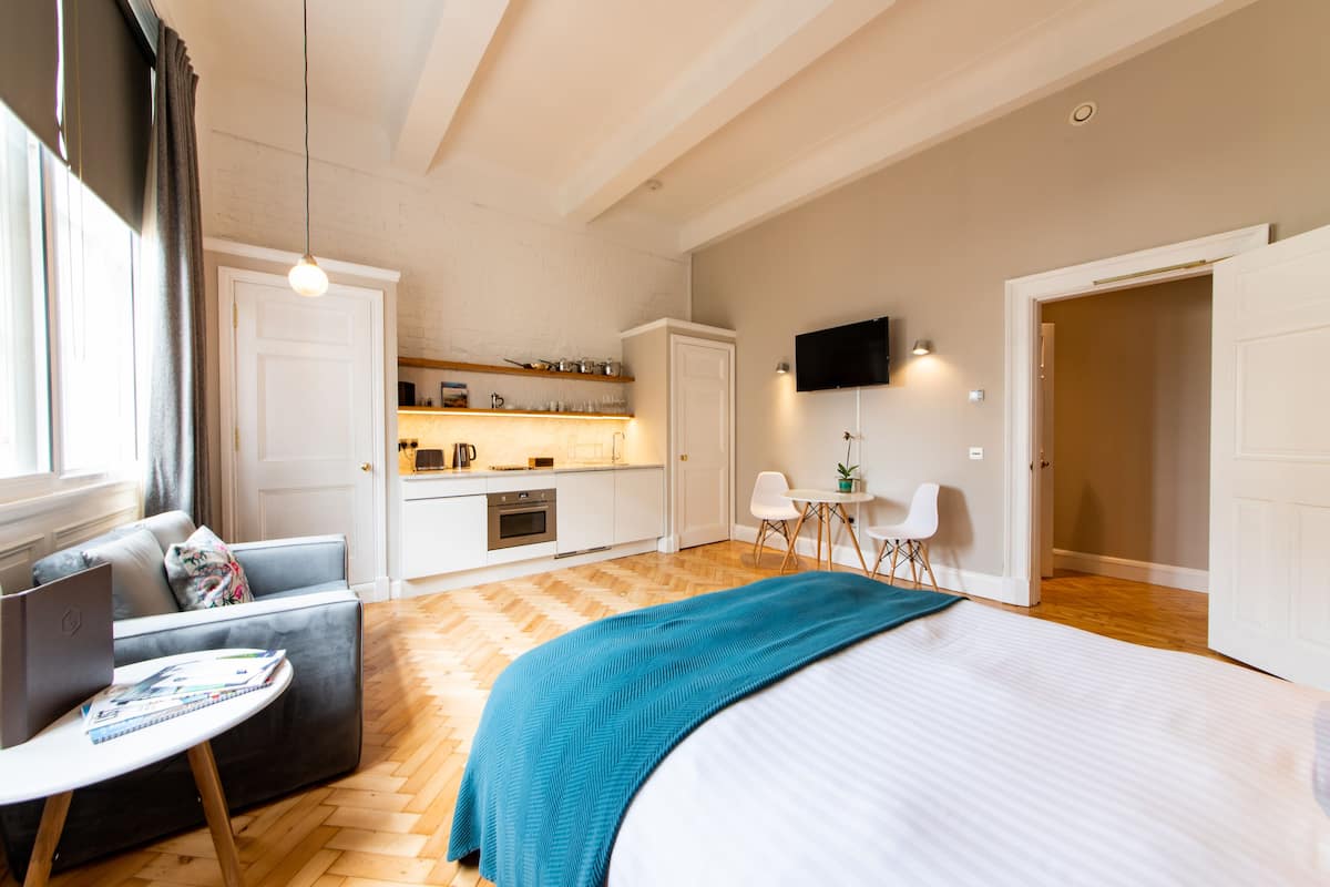 Une chambre moderne avec un grand lit, un coin salon avec une table et une chaise, une kitchenette et un téléviseur fixé au mur.