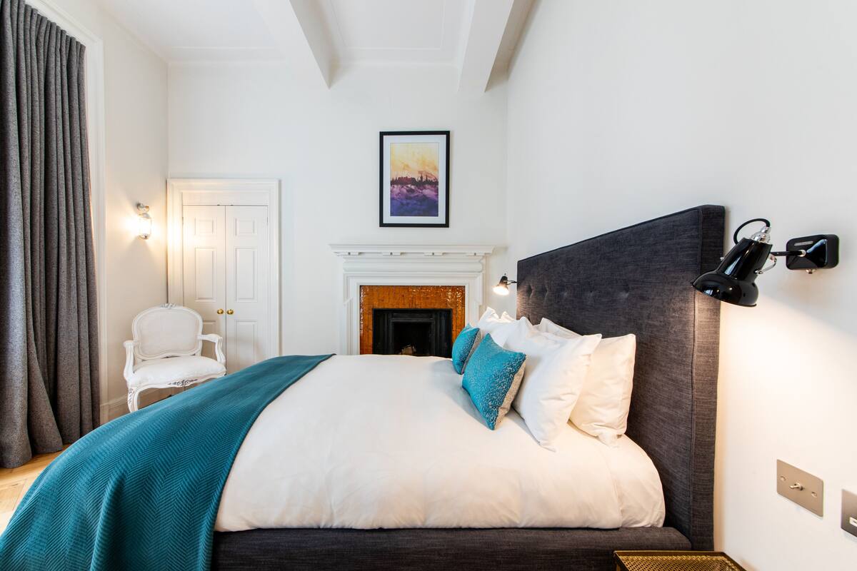 Une chambre avec un grand lit, une cheminée, un fauteuil blanc et un tableau accroché au mur.