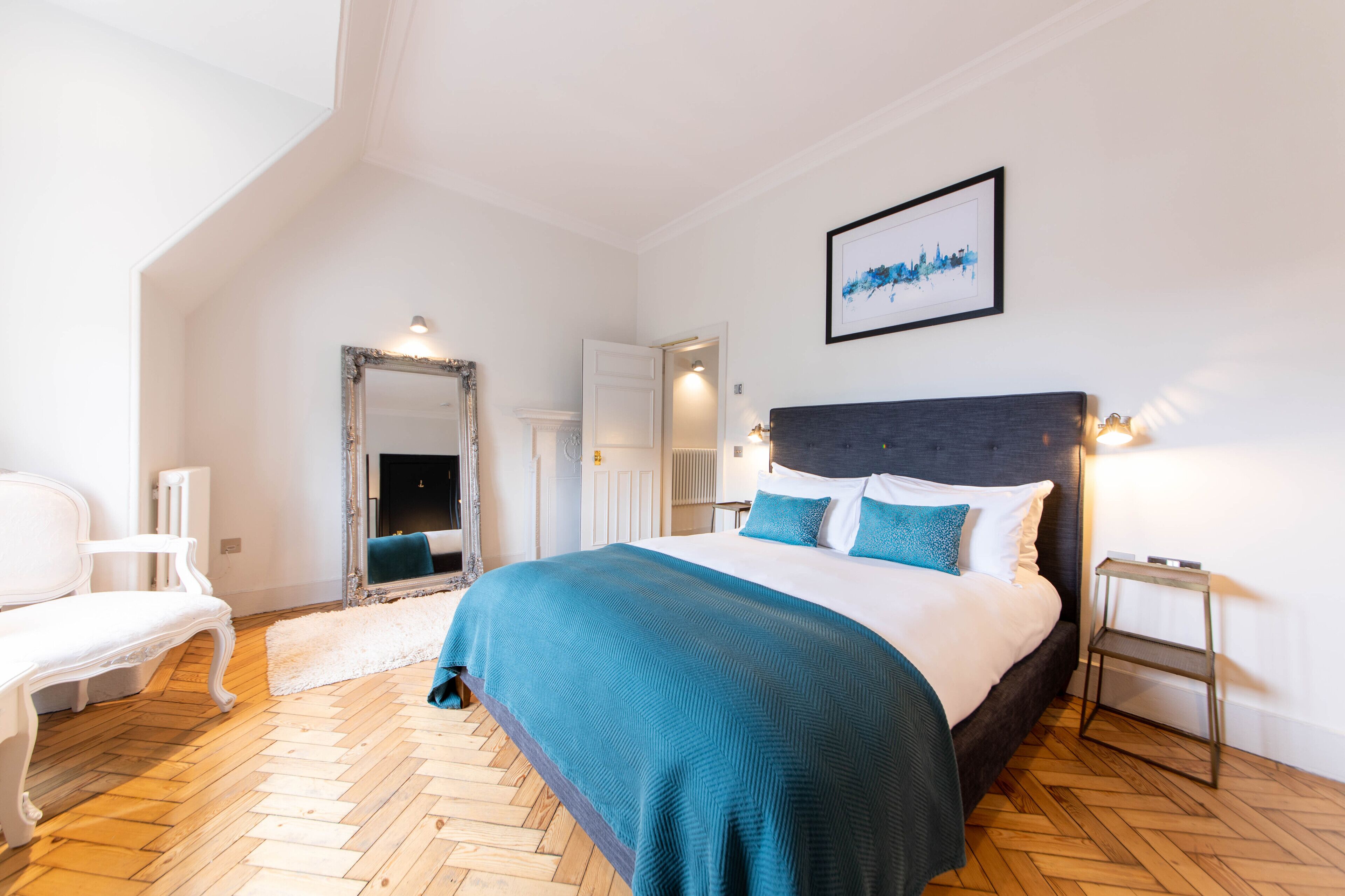 A bedroom with a large bed, a white chair, a fireplace, and a framed cityscape picture on the wall.