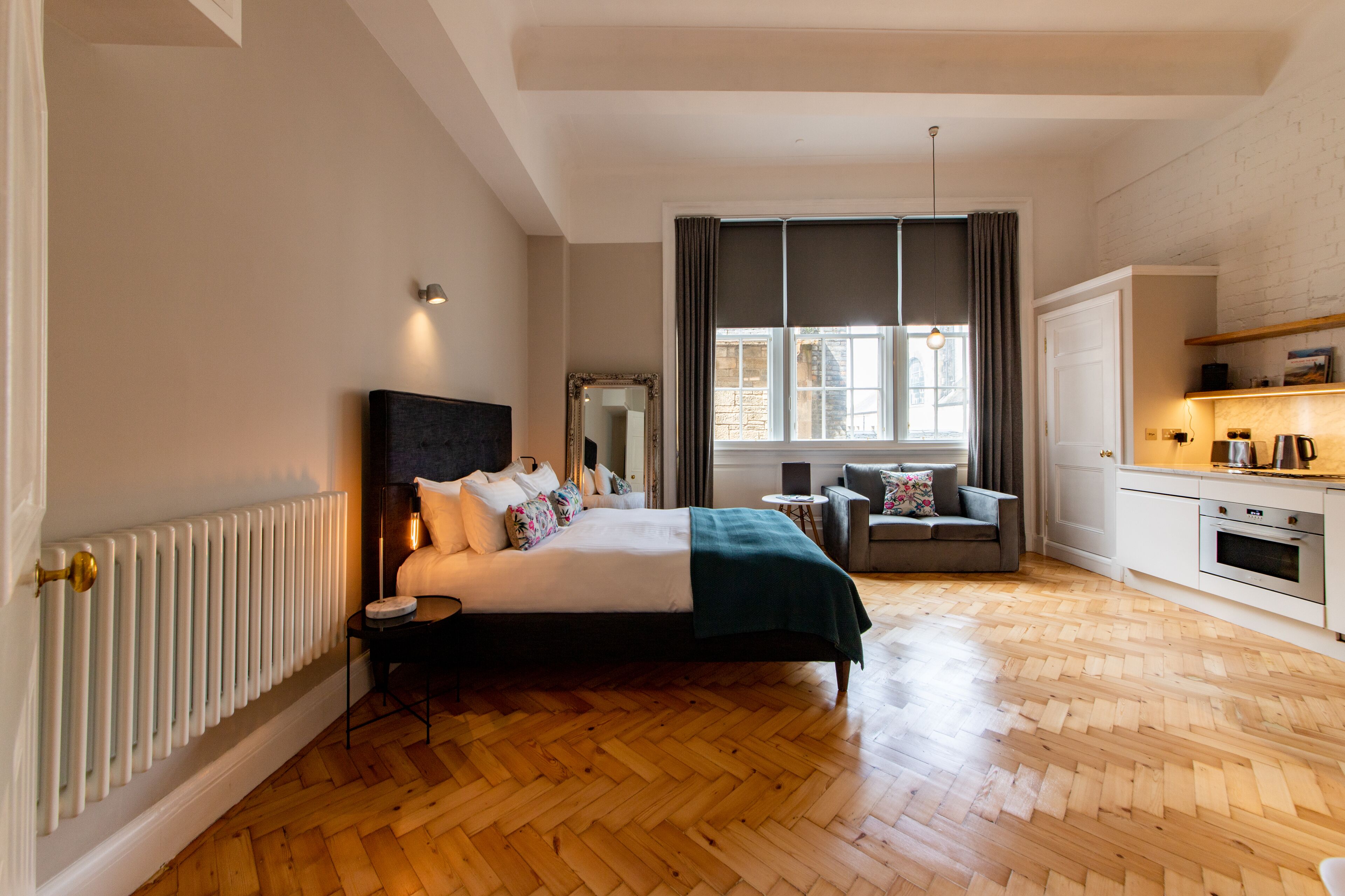 A bedroom with a large bed, a bedside table, a chair, a window with curtains, a radiator, and a kitchenette area.