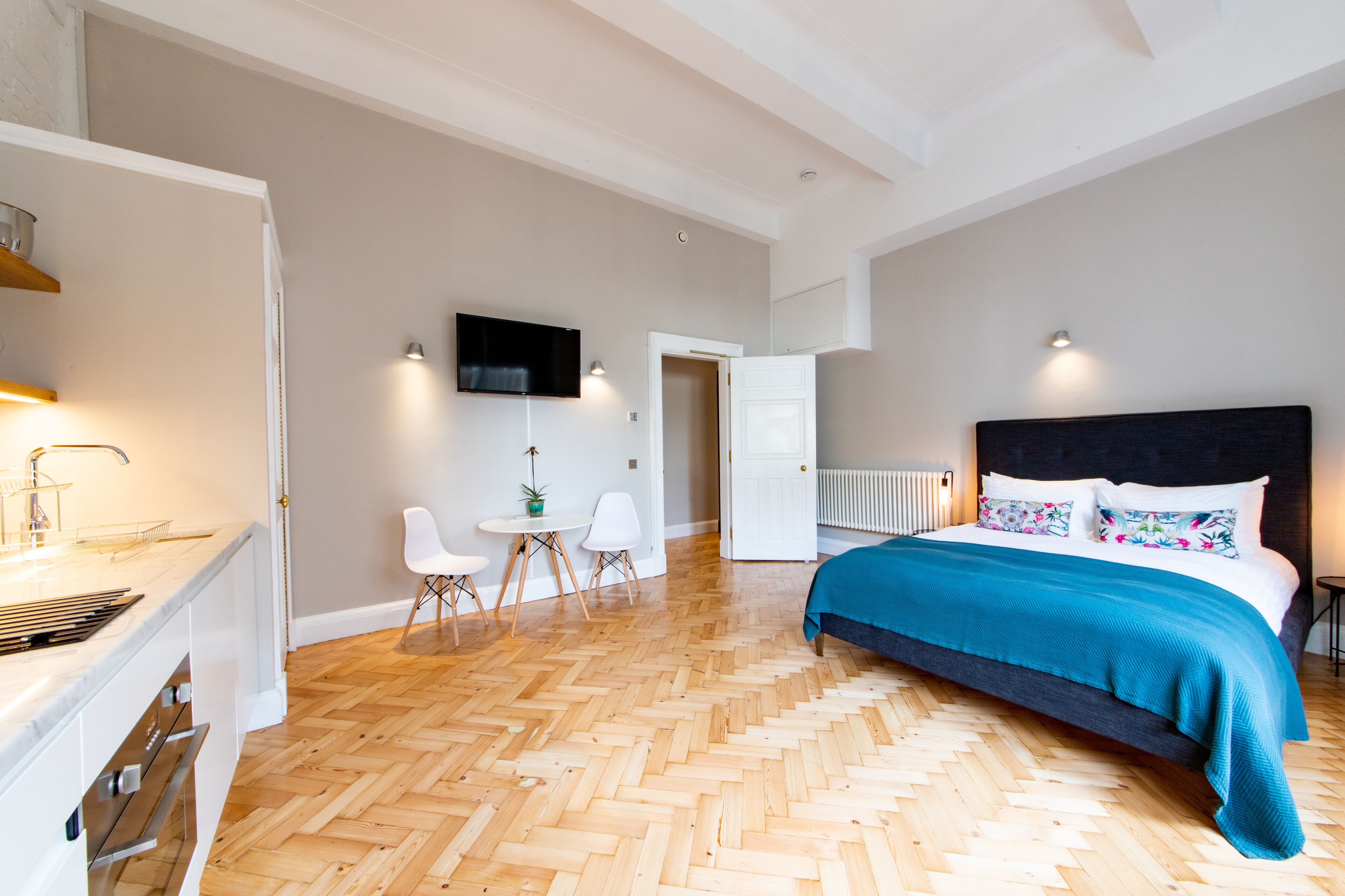 A modern bedroom with a herringbone wooden floor, a large bed with a teal bedspread, a flat-screen TV mounted on the wall, and a small dining area with a table and chairs.