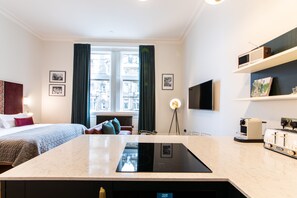 Ett modernt hotellrum med en säng, ett skrivbord, en inbyggd spis, en platt-TV och ett fönster med gardiner.