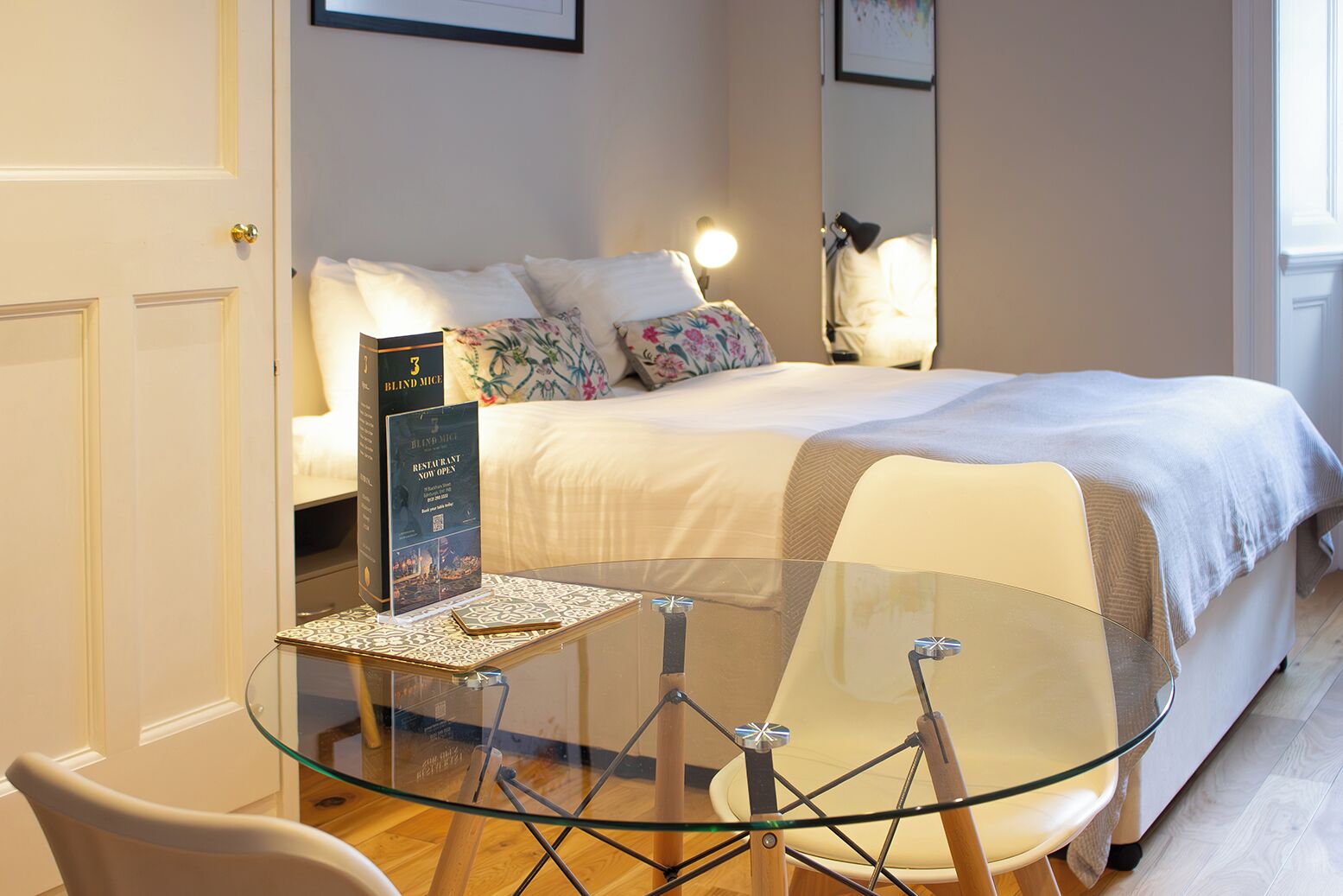 Une chambre d’hôtel avec un lit, une table en verre avec des chaises, une petite table avec un menu et une porte.
