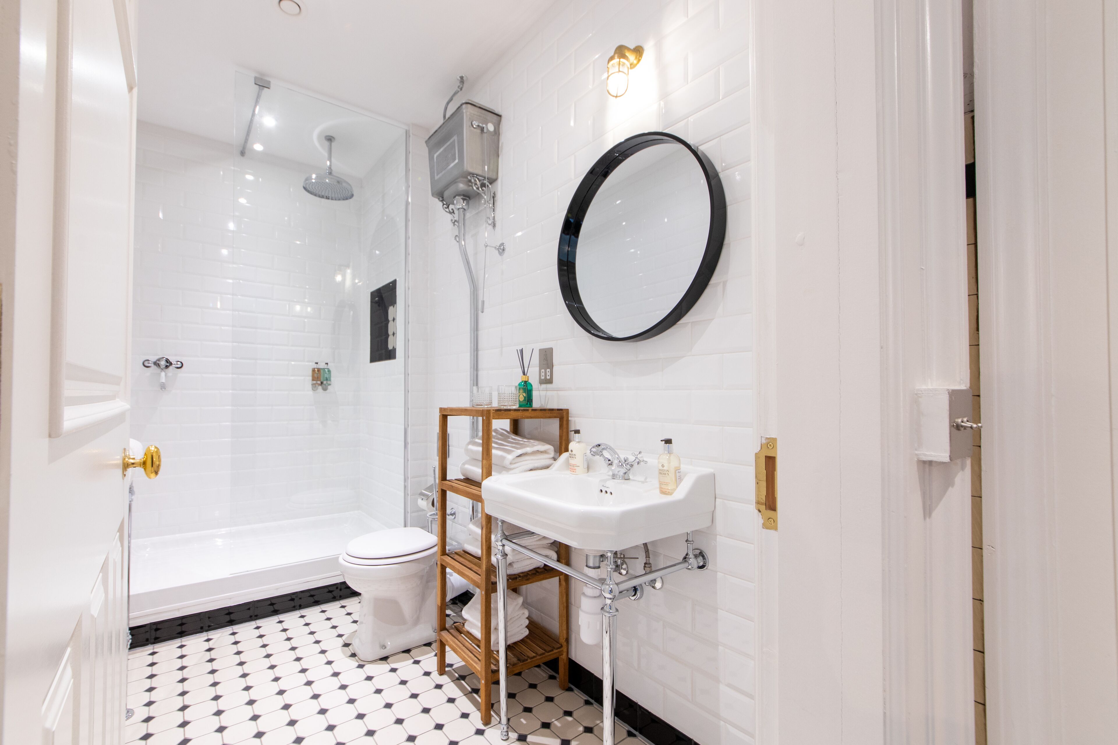 A modern bathroom with a white tiled shower, a toilet, a sink with a wooden shelf, and a round mirror.