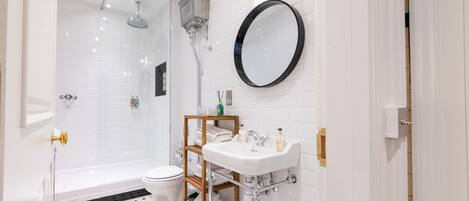 A modern bathroom with a white tiled shower, a toilet, a sink with a wooden shelf, and a round mirror.