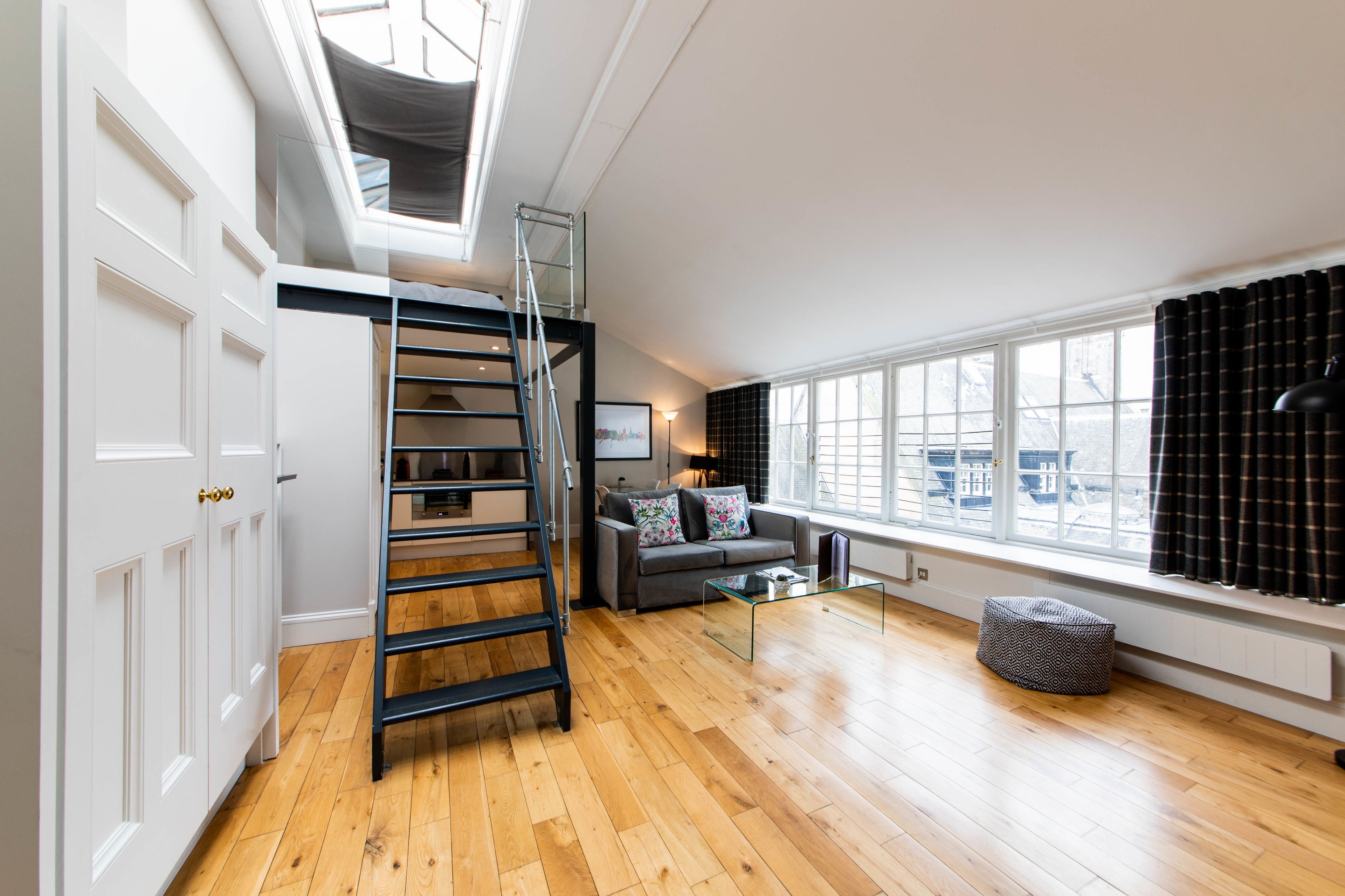 A spacious room with a wooden floor, a grey sofa, a glass coffee table, a black and white patterned ottoman, a black ladder leading to a loft, and a large window with curtains.