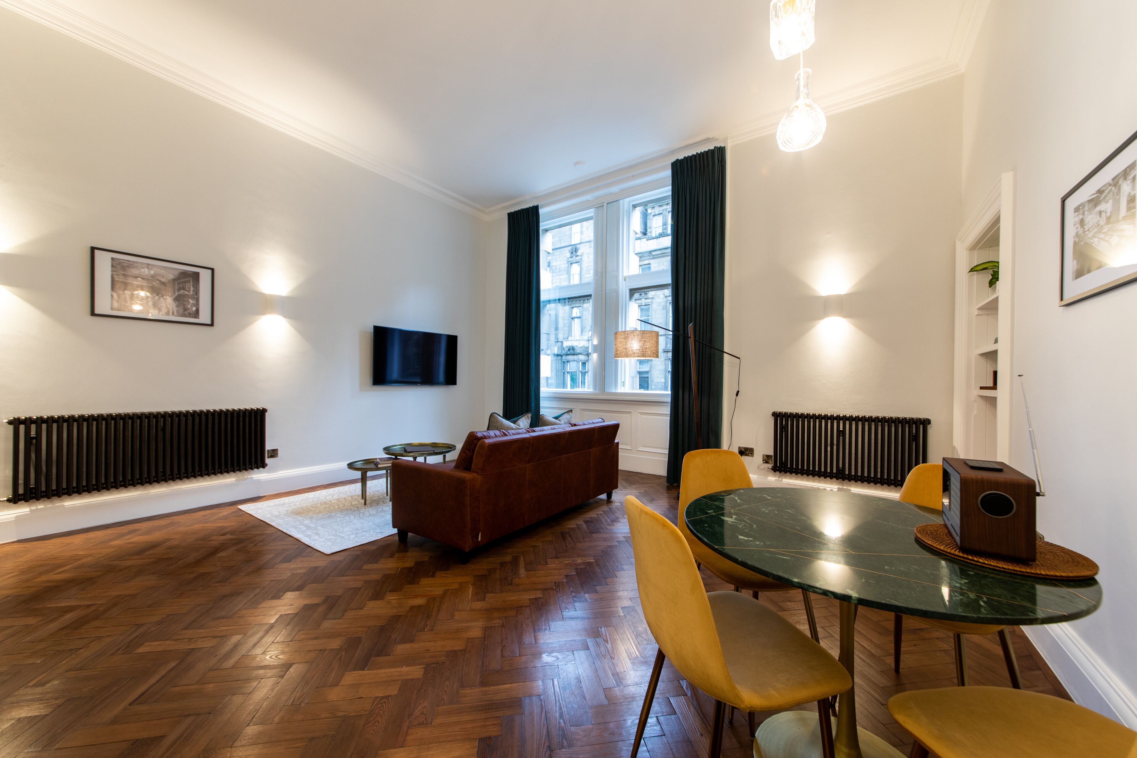 Una sala moderna con suelo de madera en patrón de espiga, un sofá de cuero marrón, una mesa de centro de vidrio y sillas amarillas. Hay un televisor de pantalla plana montado en la pared, un radiador y una ventana con cortinas.