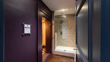 A narrow hallway with a glass shower enclosure, a wooden floor, and a door leading to a room with a tiled wall.