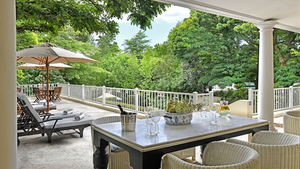 Een overdekt terras met een eettafel, rieten stoelen en een grote parasol.