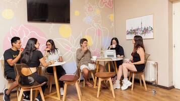 Un groupe de personnes est assis autour d’une table dans une pièce dont un mur est décoré d’une fresque florale et où est accroché un tableau représentant un paysage urbain.