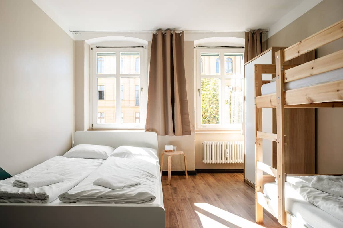 A bedroom with a bed, a wooden bunk bed, a radiator, and windows with curtains.
