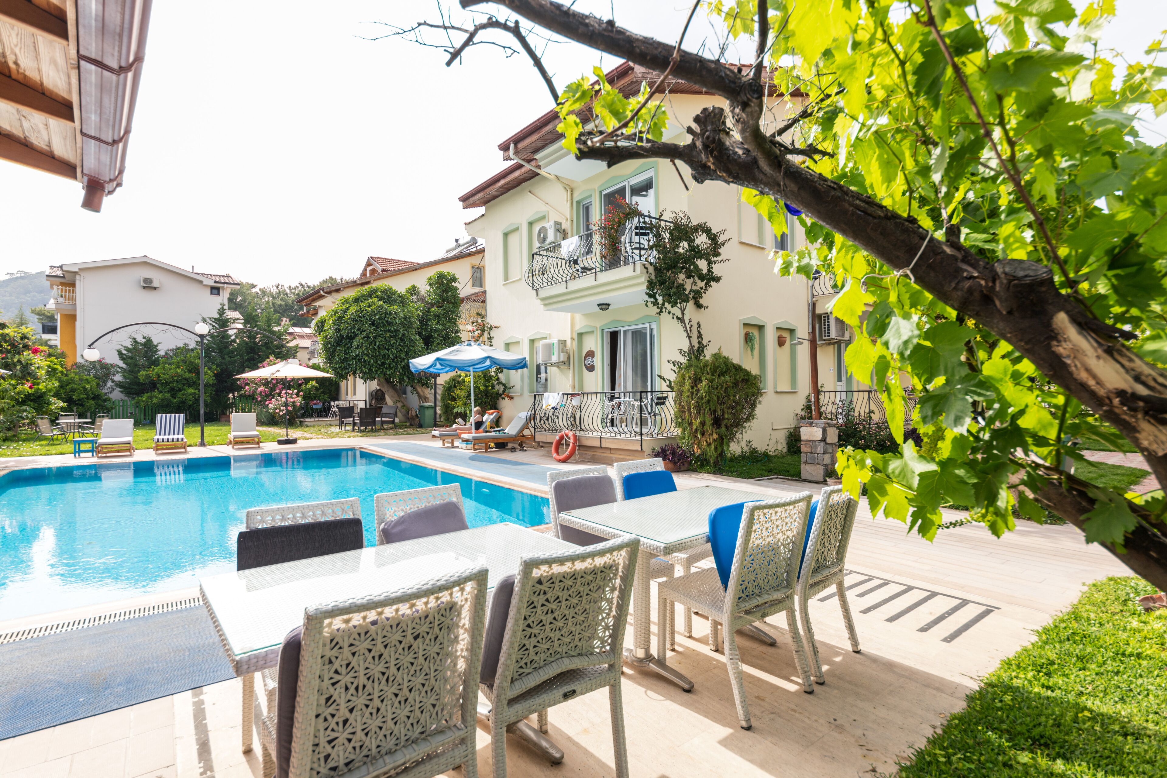 Zona de piscina con sillones de descanso y una mesa, rodeada de vegetación y edificios.