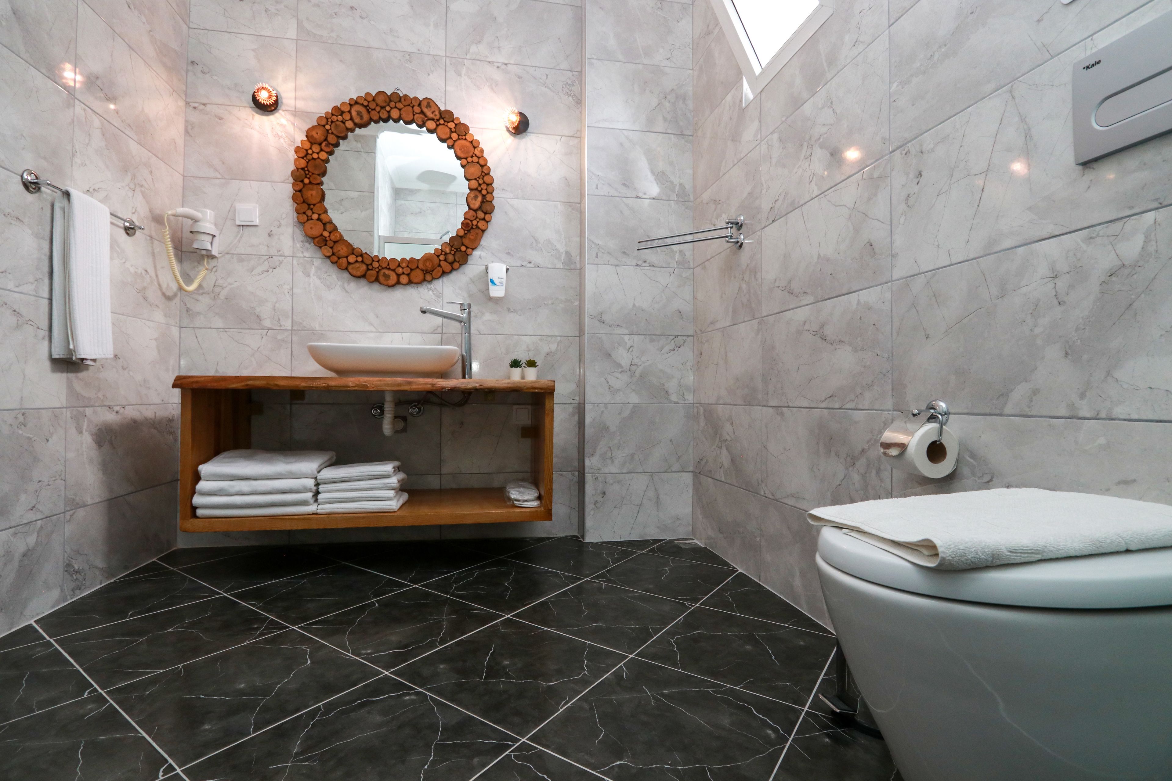 A modern bathroom with a wooden vanity, a round mirror with a wood frame, a toilet, and a towel rack.