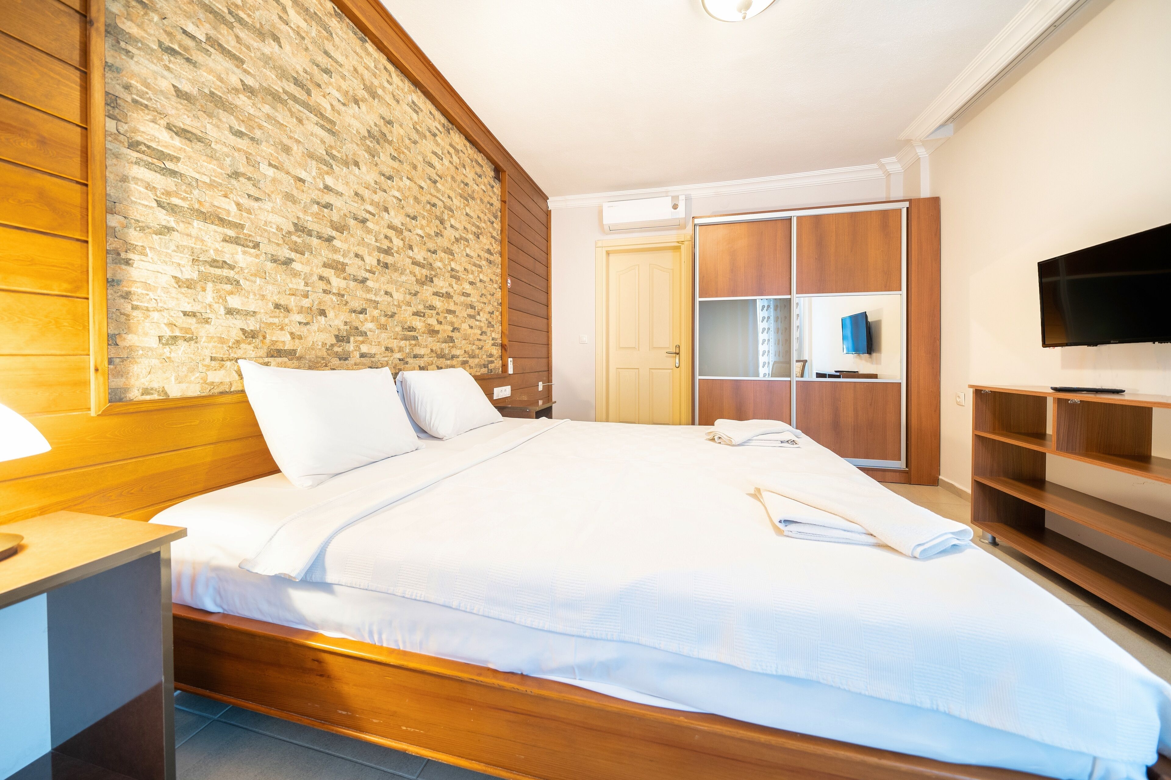 A hotel room with a large bed, wooden headboard, a brick accent wall, a wardrobe, a TV, and a desk with a lamp.