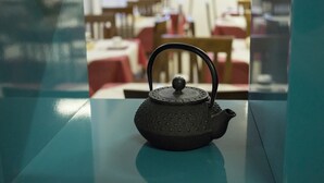 A black teapot on a teal table in a restaurant with red chairs and tables in the background. - Blue Sky (Rimini)